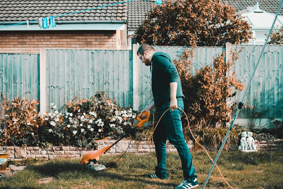 A man with short hair and glasses is performing garden maintenance in a backyard, using a yellow electric grass trimmer to cut the lawn. He is dressed in a dark hoodie, blue jeans, and striped sneaker