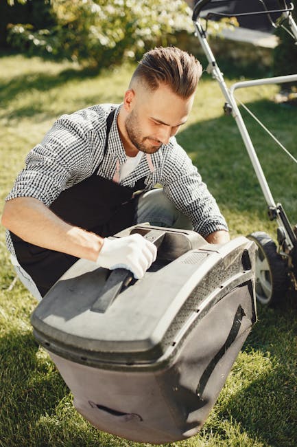 A man with short hair and glasses is performing garden maintenance in a backyard, using a yellow electric grass trimmer to cut the lawn. He is dressed in a dark hoodie, blue jeans, and striped sneaker