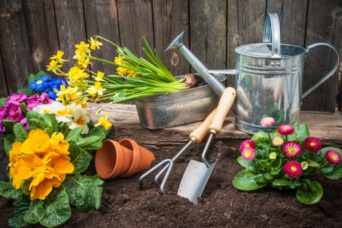 The image displays a typical gardening scene featuring various garden tools and plants. In the foreground, there is rich, dark soil with a small, empty terracotta plant pot placed on its side. Surrounding the soil are colorful flowering plants, including a vibrant yellow primula, pink and white begonias, and purple pansies, all with healthy green foliage. Behind the flowers, a pair of gardening tools—a hand fork and a hand trowel with wooden handles—are partially inserted into the soil, indicating ongoing planting or garden maintenance. To the right of the tools, a silver metal watering can is positioned, with its spout directed downward. Near the watering can, two terracotta pots are stacked, ready for use. In the background, a wooden garden fence provides a neutral backdrop, emphasizing the natural outdoor environment. The scene is well-lit by natural daylight, suggesting a clear weather day suitable for gardening activities. This setup illustrates typical gardening preparations and care, especially relevant for services such as lawn and plant bed maintenance offered by Gardener Yiewsley, located near Yiewsley, Middlesex, with a focus on enhancing outdoor spaces in the local area.